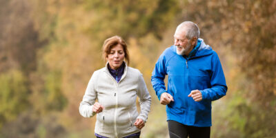 An older couple jogging through a forest in autumn